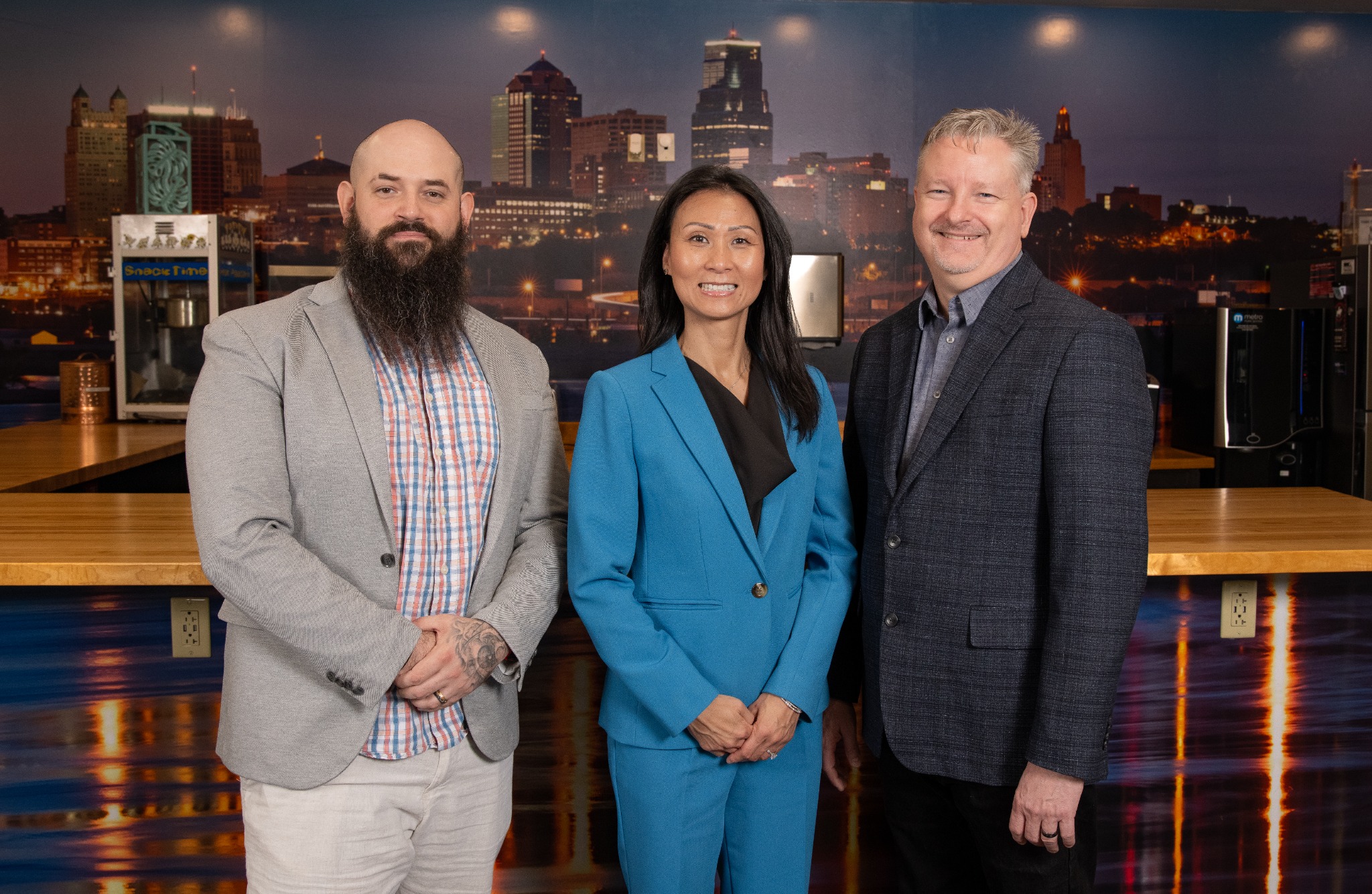 Three professionals standing in front of a city skyline backdrop in a modern office setting, smiling at the camera.